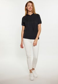 Sweat-shirt à manches courtes noir avec cordons, associé à un pantalon blanc ajusté et des baskets blanches, se tenant devant un fond clair.