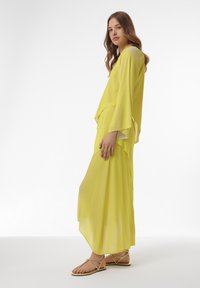 Geel gelaagd outfit van lichtgewicht stof, met ruime mouwen en een vloeiende rok, aangevuld met strap sandalen.