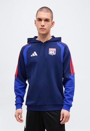 Jeune homme portant un sweat à capuche bleu marine Olympique Lyonnais avec des bandes rouges sur les manches et un short noir, debout devant un fond uni.