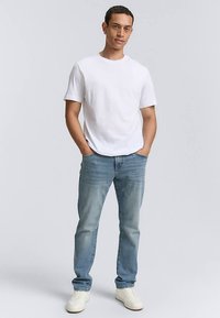 T-shirt en coton blanc, jean denim bleu clair et baskets blanches portés par une personne debout. Design simple, coupe décontractée, sans motifs ni logos.