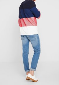 Crewneck, gekleurde blokken trui in marineblauw, rood en wit. Een paar lichtblauwe jeans en witte sneakers met een beige zool. Ontspannen pasvorm.