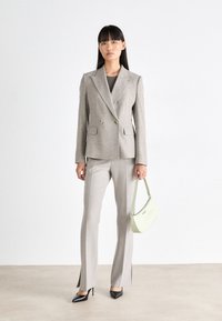 Lys grå dobbeltradet blazer med notched lapels, kombinert med matchende bukser med sidespalter. Modellen bærer en lys grønn håndveske.
