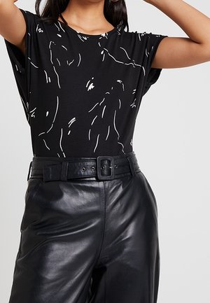 Nainen pukeutuneena mustaan t-paitaan, jossa valkoisia abstrakteja viivoja, laitettuna korkeavyötäröisiin mustiin nahkahousuihin, joissa leveä vyö ja suorakaiteenmuotoinen solki.