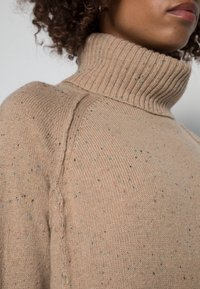 Beige kauluspaita, joka on valmistettu tekstuurillisesti neulotusta kankaasta, jossa on värikkäitä täpliä. Sisältää ribbinecktassun ja sauman yksityiskohtia hartioiden kohdalla.