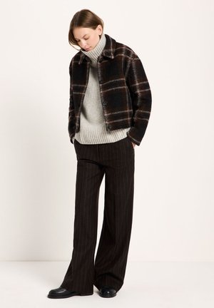 Woman wearing a black and brown plaid jacket, gray turtleneck sweater, black pinstripe wide-leg pants, and black shoes, standing with hands in pockets.
