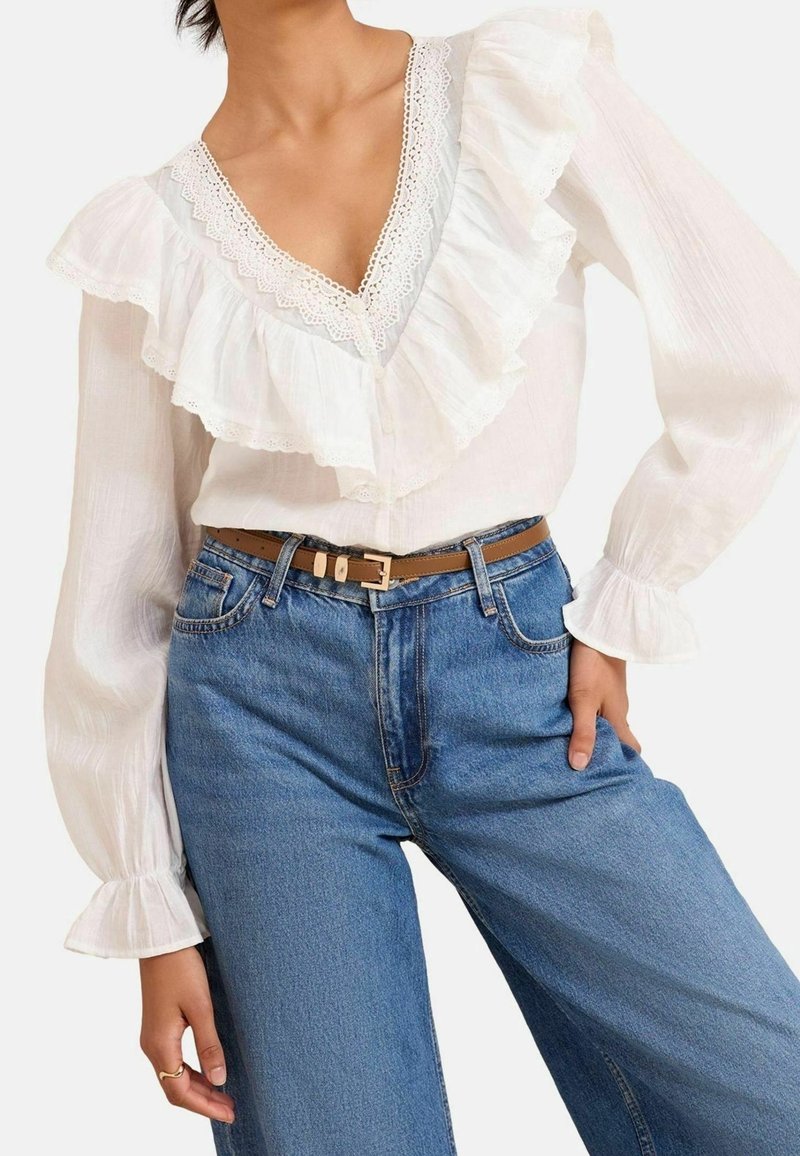 Femme portant un chemisier blanc à manches longues avec des volants en dentelle et un jean bleu taille haute avec une ceinture marron, posant avec une main sur la hanche.