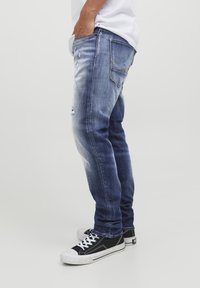 Jeans en denim bleu avec une coupe slim, présentant des détails de déchirure et une légère décoloration. Associés à des baskets basses noir et blanc.