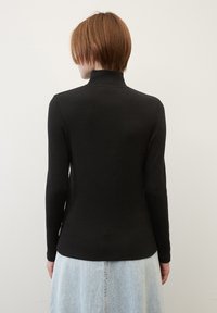 Persona con capelli corti castano chiaro sta in piedi di spalle, indossando un maglione a collo alto nero a maniche lunghe e una gonna in denim blu chiaro, davanti a una parete bianca.