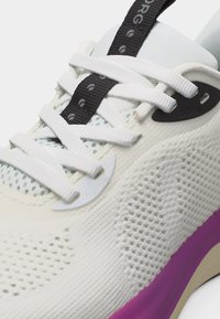 Κοντινό πλάνο λευκού πλεκτού αθλητικού παπουτσιού με λευκά κορδόνια, μαύρο λουράκι στην γλώσσα και μοβ διακριτικό στη σόλα.