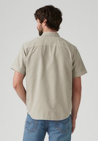 Short-sleeve, collared shirt in light beige fabric with a smooth texture. Features stitching details along the back and a curved hem.