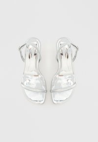 Ασημί ψηλοτάκουνες σανδάλια με γυαλιστερή φινέτσα, που διαθέτουν δύο ρυθμιζόμενες λωρίδες και ασφαλή κούμπωμα στον αστράγαλο, λεία επιφάνεια και ανοιχτό σχέδιο.