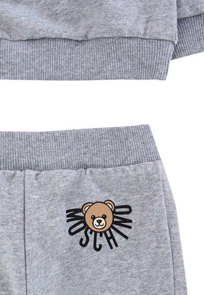 Szary dres z bawełny z domieszką włókien, z ściśniętymi mankietami, grafiką misia oraz napisem "Moschino" na spodniach. Miękka faktura i luźny krój.