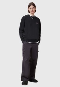 Felpa nera con design a forma di cuore, abbinata a pantaloni cargo scuri. L'outfit presenta una vestibilità rilassata e un design minimalista, enfatizzando il comfort.