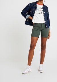 Chaqueta de mezclilla, camiseta blanca con texto, pantalones cortos verdes oliva, zapatillas blancas de caña alta. Presenta mangas enrolladas y un corte casual.