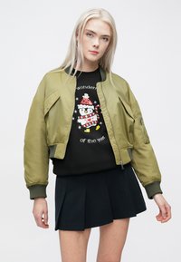 Olive zaļš bomber jakas mūžīgām piedurknēm, kopā ar melnu sporta krekla, kurā ir pingvīna dizains, un īsu melnu svārku ar krokām.