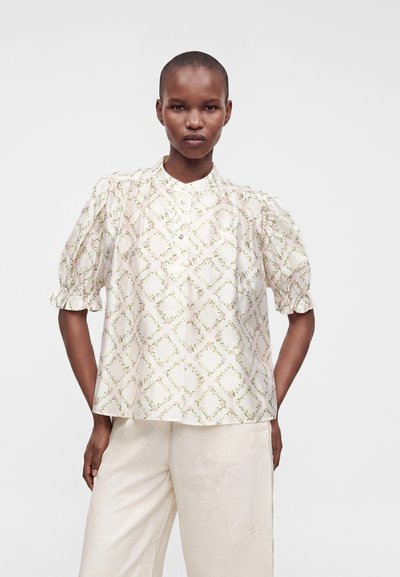 Femme aux cheveux courts porte un chemisier blanc avec motif floral en losange et manches bouffantes, associé à un pantalon blanc ample.