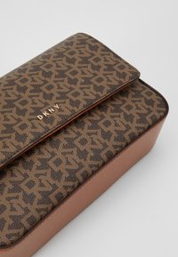 Hnědá peněženka DKNY s tmavohnědým monogramem a zlatým logem na texturovaném koženém povrchu, na bílém pozadí.