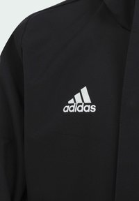 Černá bunda vyrobená z hladkého materiálu s bílým logem Adidas na hrudi. Design zahrnuje vysoký límec a zipový uzávěr.