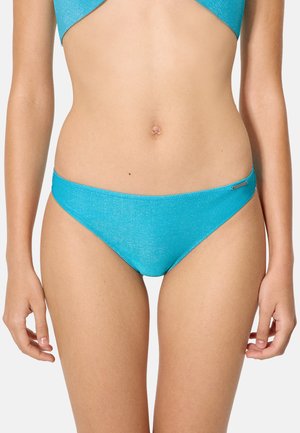 Mujer con bikini turquesa brillante de abajo y arriba a juego, de pie con los brazos relajados a los lados contra un fondo liso.