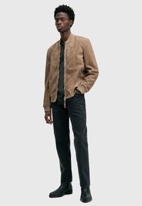 Giubbotto bomber in pelle beige con chiusura a zip e tasche laterali, indossato sopra una maglietta nera, abbinato a jeans neri e stivali neri alla caviglia.