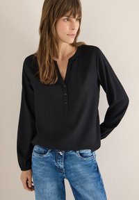 Blouse noire à manches longues avec un col en V et une patte de boutonnage, portée avec un jean en denim bleu clair. Tissu lisse, coupe décontractée, avec des poignets froncés.