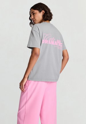 Femme aux cheveux courts et bouclés portant un t-shirt gris avec un texte rose "Mignonne mais dramatique" au dos et un pantalon rose ample, debout de profil.