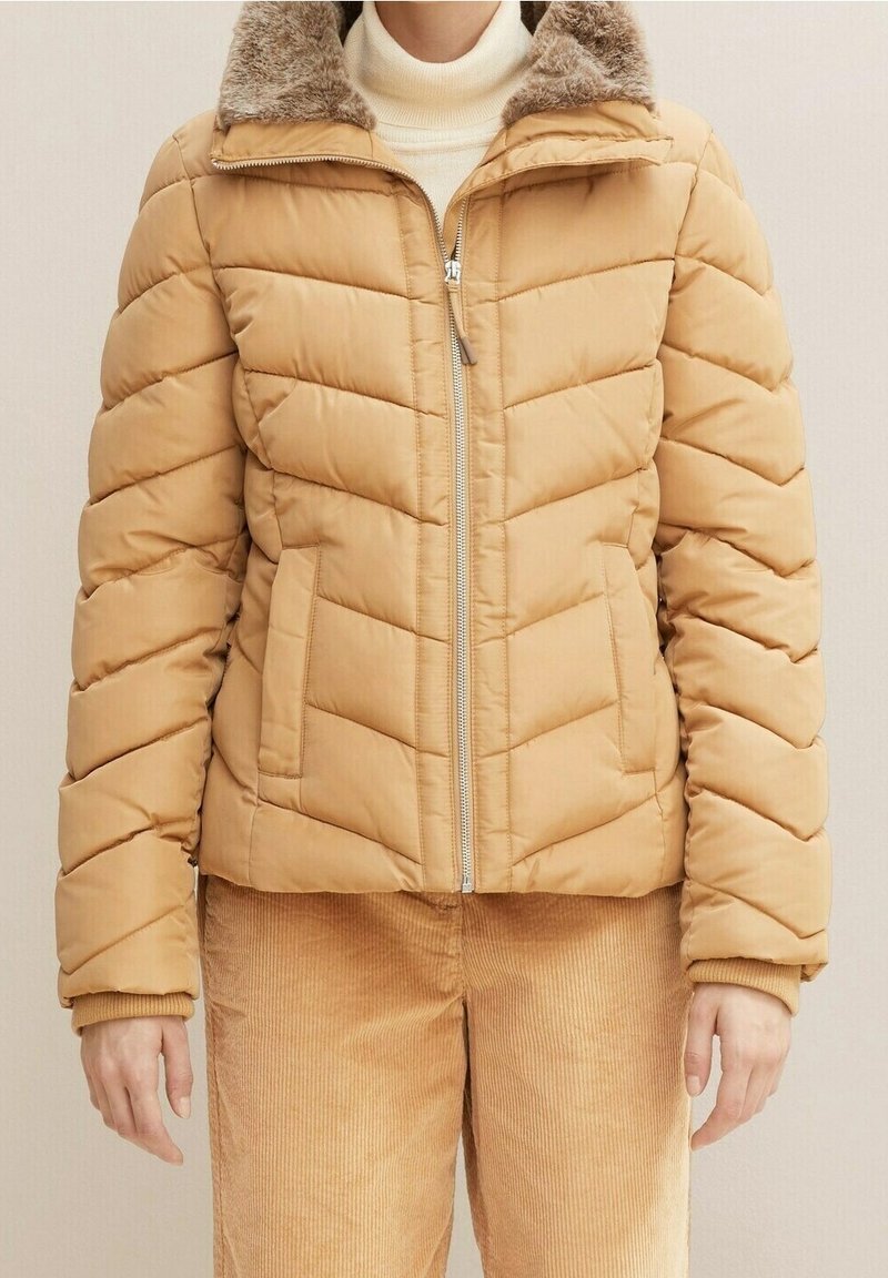Personne portant une veste matelassée beige avec un col en fourrure et un pantalon en velours côtelé beige, debout devant un fond uni.