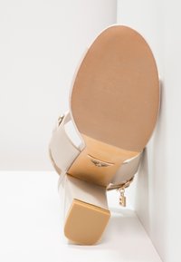Sandale blanche à talon haut avec semelle texturée, talon carré, bride à la cheville et breloque boucle dorée, positionnée semelle vers le haut contre les murs d'un angle.