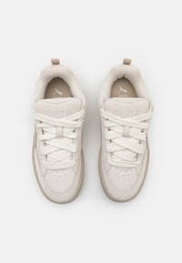 Sapatilhas de cor creme com parte superior em camurça e malha, atacadores grossos, sobreposições texturizadas e sola de borracha. Logo "PUMA" na língua.
