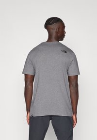 Camiseta de algodón gris con mangas cortas, cuello redondo y el logo de The North Face en negro en la parte superior de la espalda. Tela texturizada y corte relajado.