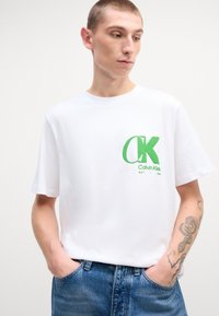 Camiseta de algodón blanca con un gran logo verde de "CK" y el texto "Calvin Klein". Manga corta, corte relajado, combinada con jeans azules.