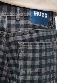 Κοντινή λήψη στην τσέπη καρό παντελονιού σε αποχρώσεις μπλε και γκρι με ραμμένη μπλε ετικέτα της μάρκας "HUGO" πάνω από την τσέπη.