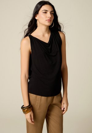 Donna con lunghi capelli scuri che indossa una canotta nera, pantaloni beige, grandi orecchini a cerchio e braccialetti grossi a un polso.