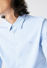 Camicia a righe blu chiaro con colletto abbottonato, bottoni bianchi e un piccolo logo ricamato sul petto. Tessuto di texture liscia.