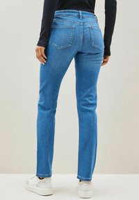 Blue denim jeans with a slim fit, mid-rise waist, and five-pocket design. Texture is smooth with a slight fade. Paired with white sneakers.