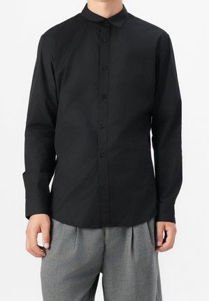 Mężczyzna ubrany w dopasowaną czarną koszulę na guziki z długim rękawem i kołnierzykiem oraz szare plisowane spodnie na jednolitym tle.