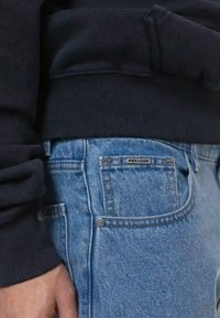 Jeans de denim em azul claro com uma textura suave. Apresenta um bolso traseiro etiquetado, botão de metal prateado e um sweatshirt preto por cima.