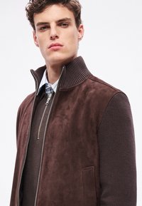 Jachetă maro din piele intoarsă cu mâneci tricotate cu textură, guler înalt și fermoar frontal. Dispune de buzunare laterale și un finisaj neted.