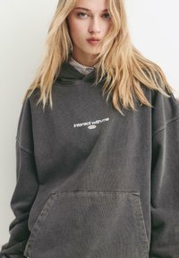 Grauer übergroßer Hoodie mit dem Text "interagiere mit mir". Verfügt über eine Kängurutasche, gerippte Bündchen und einen weichen, strukturierten Stoff.