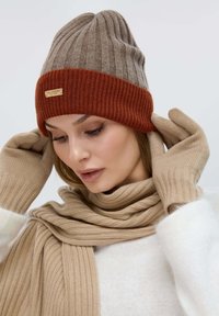 Cappello a berretto in marrone e ruggine, con texture a coste e un'etichetta con logo. Guanti e sciarpa beige con un motivo a coste simile completano il look.