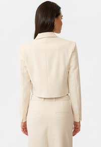 Blazer beige court avec col à revers, coupe ajustée, manches longues et subtil éclat. Comprend deux fentes à l'arrière et des accents de boutons sur les poignets.