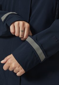 Chaqueta impermeable de color marinero con una banda reflectante gris en el puño y un bolsillo con cremallera, fabricada con un tejido liso y duradero.