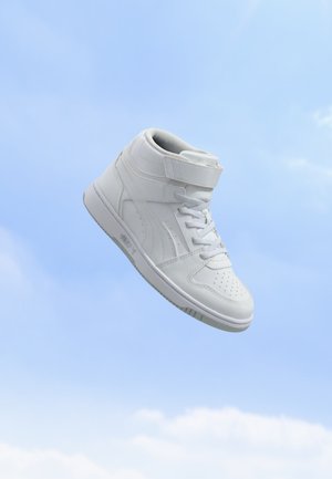 Baskets montantes blanches avec lacets et bande velcro flottant sur fond de ciel bleu.