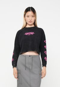 Tricou negru scurt, cu mânecă lungă, având un logo grafic roz pe piept și modele cu cranii pe mâneci. Purtați cu o fustă cargo gri.