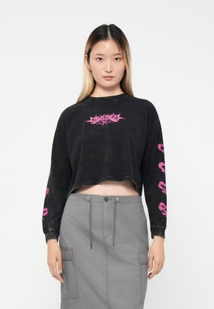 Cropped black long-sleeve shirt with a pink graphic logo on the chest and skull patterns on the sleeves. Worn with a grey cargo skirt.