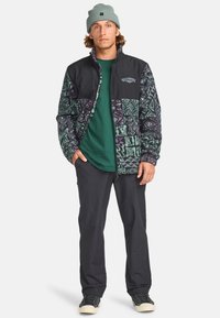 Veste en polaire avec partie supérieure noire et partie inférieure à motifs verts et violets. Associée avec une chemise verte et un pantalon sombre, portée avec des baskets.