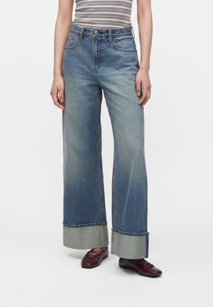 Persoon draagt een blauwe jeans met hoge taille en wijde pijpen met grote omslagen, gecombineerd met bordeauxrode veterschoentjes en een mouwloos gestreept topje.