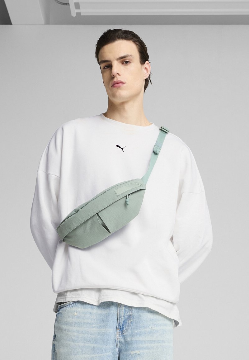 Marsupio verde chiaro con una superficie liscia, due tasche frontali e cinturino regolabile. Indossato sopra un sudario bianco e jeans blu.