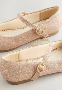 Bailarinas brillantes en color rosa con una tira, que presentan una hebilla decorativa dorada. Forro de tela suave con un patrón de flores en la plantilla.