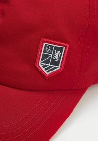 Czerwona czapka z naszytym emblematem przedstawiającym lwa oraz geometryczny wzór w czerni i bieli na tarczowym łaty. Gładka faktura materiału.